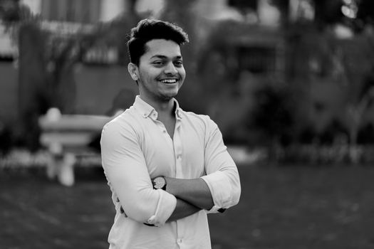 Black and white of young cheerful guy in classic shirt standing with crossed arms in park and smiling